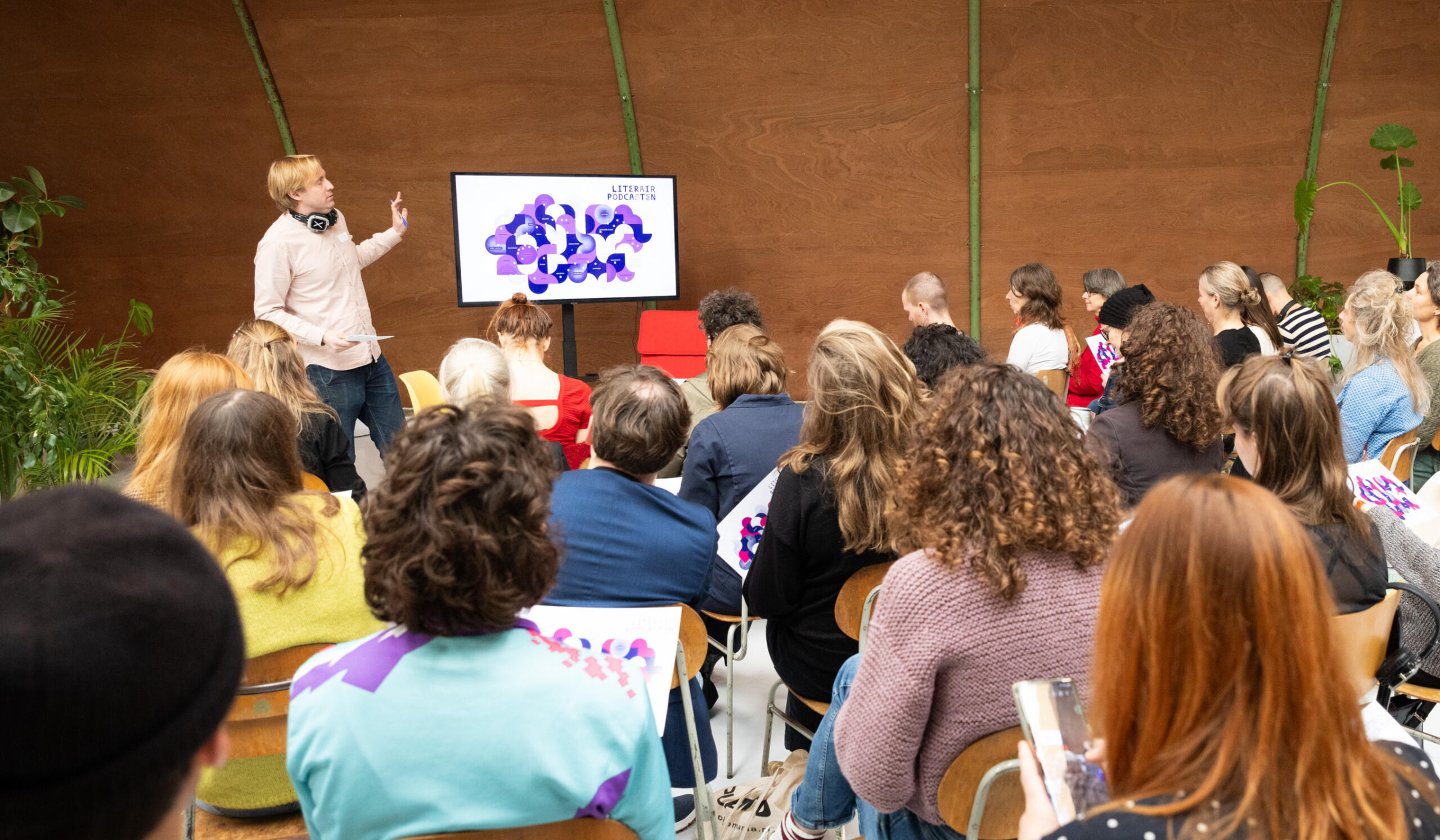 Lieven Heeremans voor een zaal met mensen die op stoelen zitten. Op het scherm staat een kaart met veel kleuren en daarin de tekst "Literair Podcasten". Foto door Lilian van Rooij